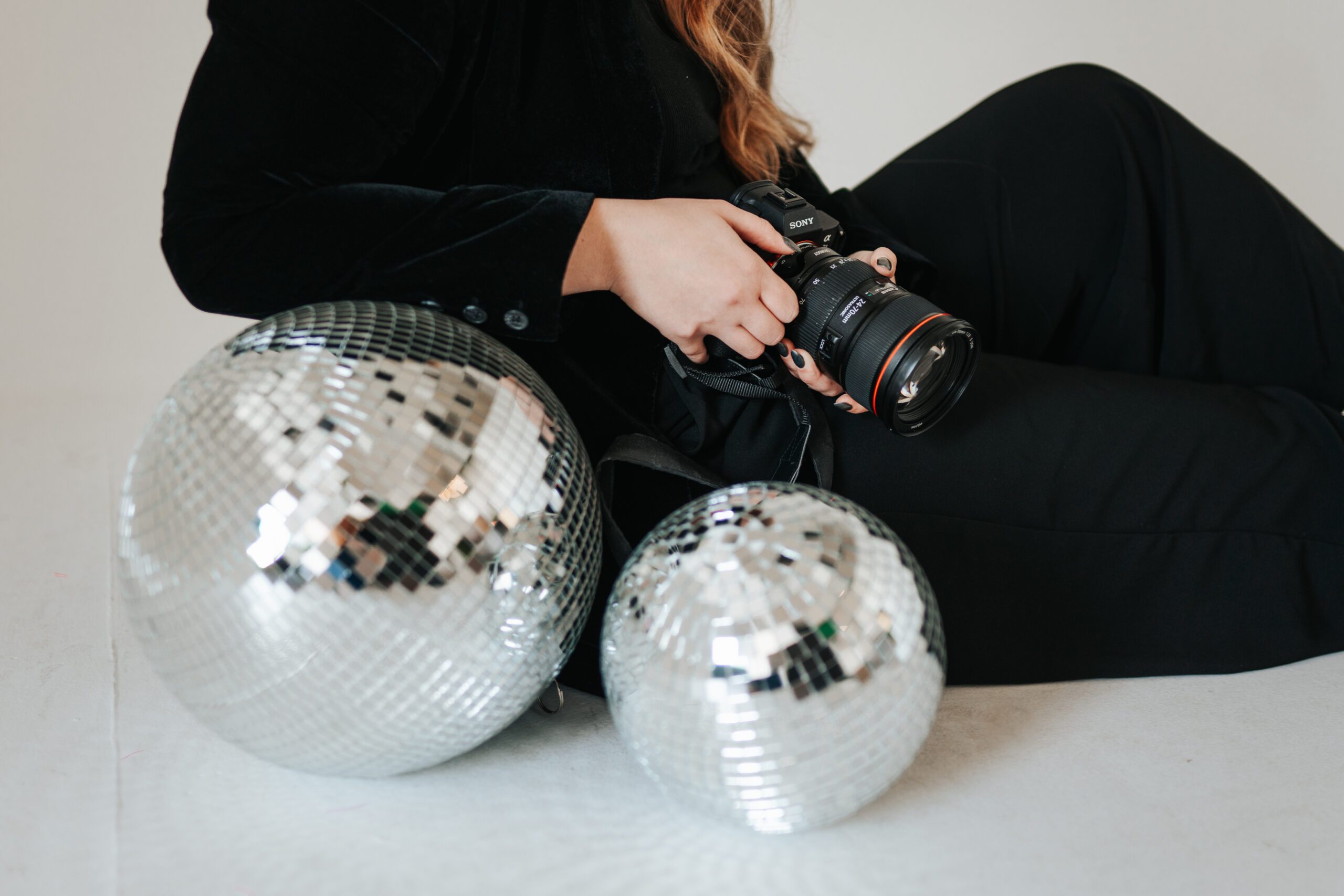 Wedding photographer sitting against white backdrop holding a camera with disco balls in the foreground