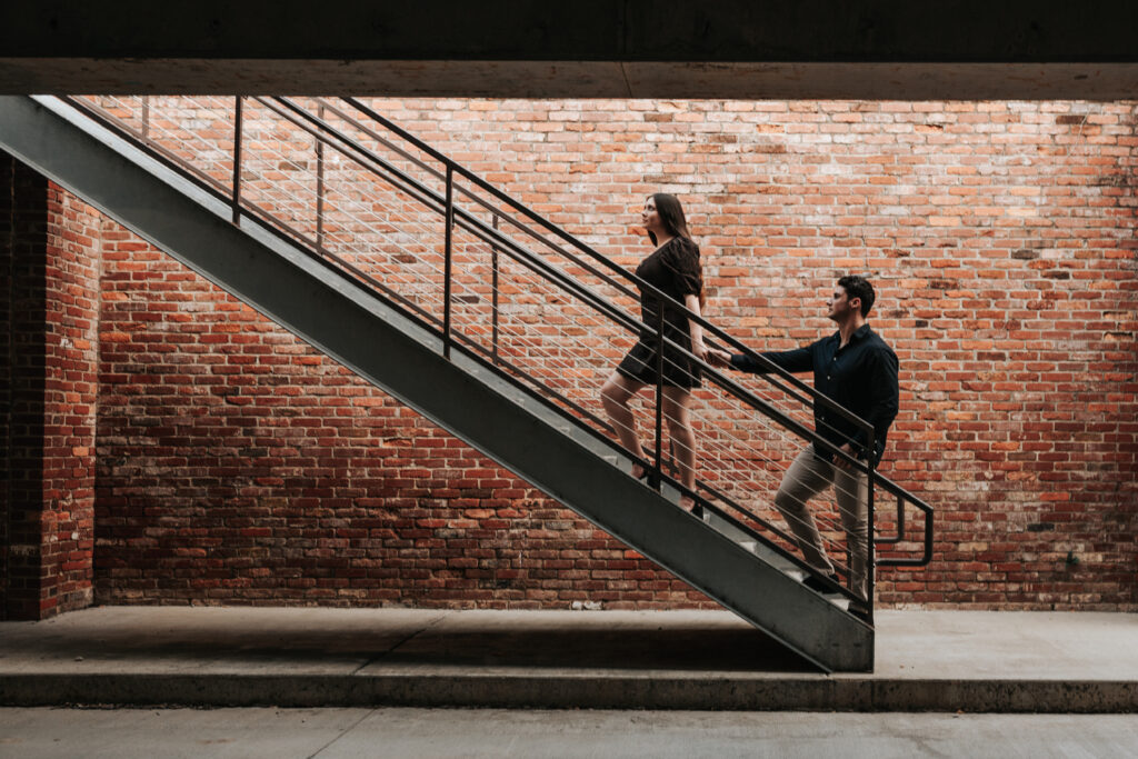 Cinematic and candid engagement photo of a couple walking up stairs in an urban setting. There are deep shadows framing the couple at the bottom of the staircase.