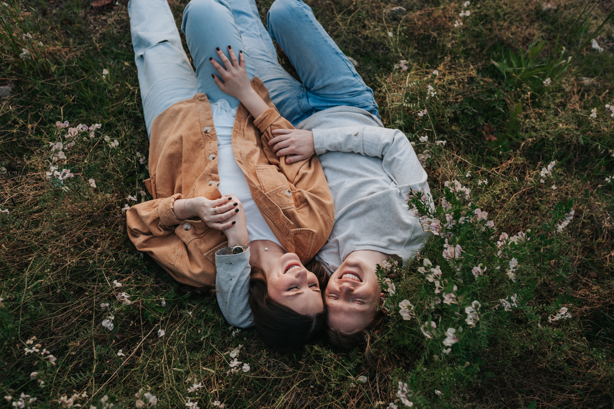 Cinematic and candid photo of a couple laying on their backs in a field of flowers. The couple is laughing and looking as the clouds go by. 