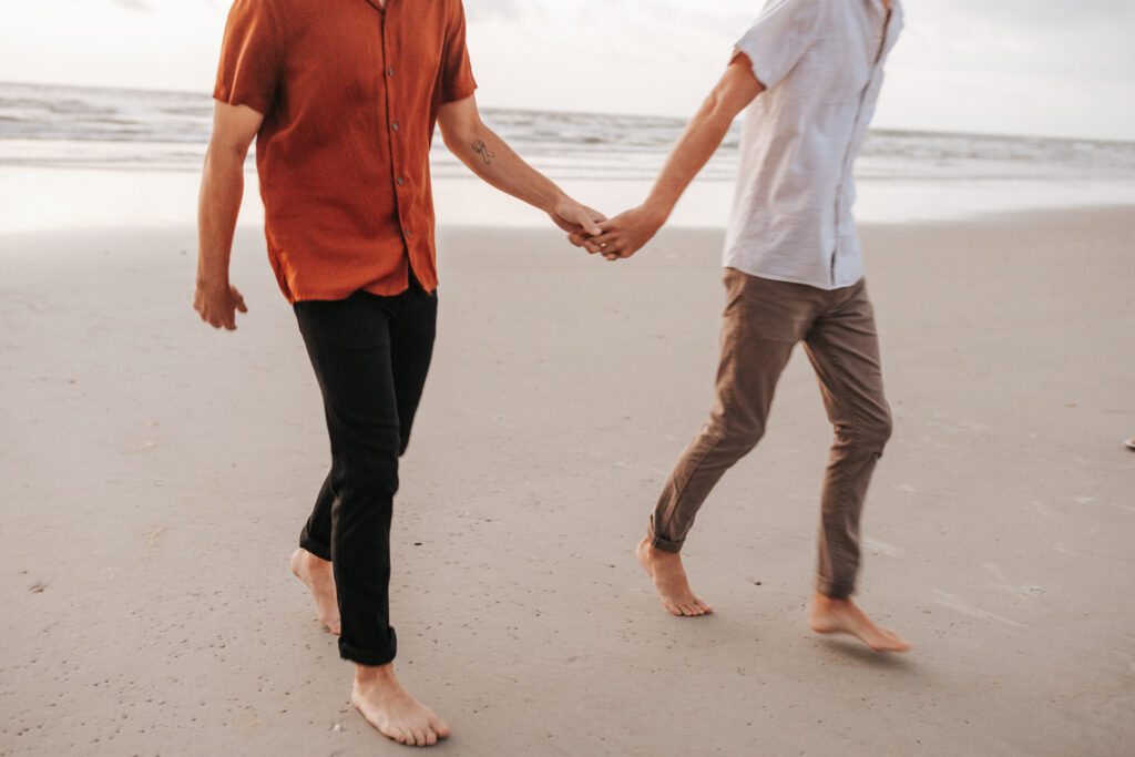 Cinematic and slightly blurry candid photo of gay couple walking hand in hand along the beach. 