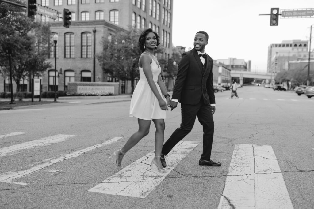 Cinematic and Candid photo of newly engaged couple crossing the street in downtown Columbus. The young couple is wearing a white dress and a black suit. 